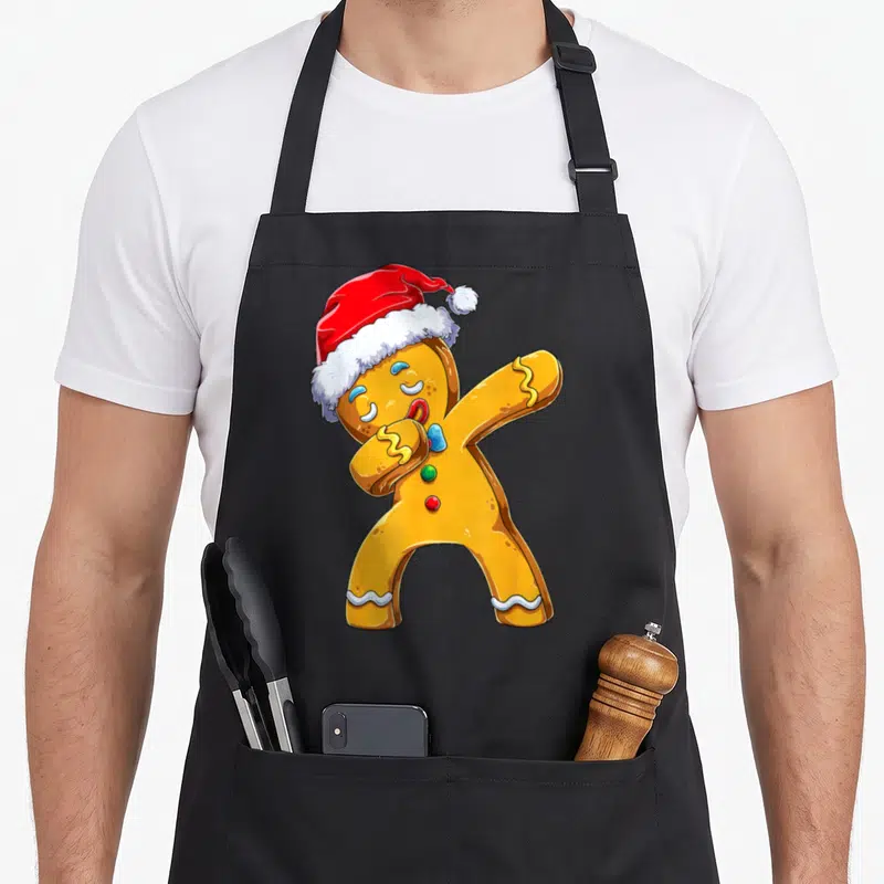 Tablier de cuisine noir avec un biscuit de Noël doré portant un bonnet rouge et effectuant une pose dab.