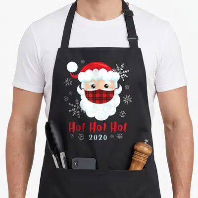 Tablier de cuisine noir avec illustration d'un Père Noël portant un masque à carreaux rouge et blanc entouré de flocons de neige.