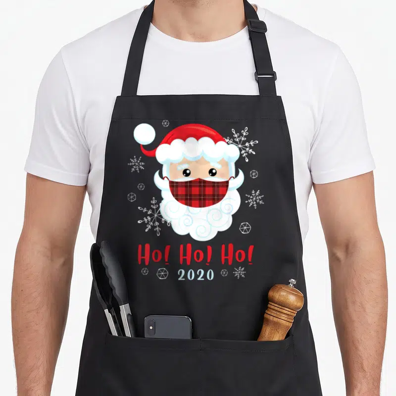 Tablier de cuisine noir avec illustration d'un Père Noël portant un masque à carreaux rouge et blanc entouré de flocons de neige.