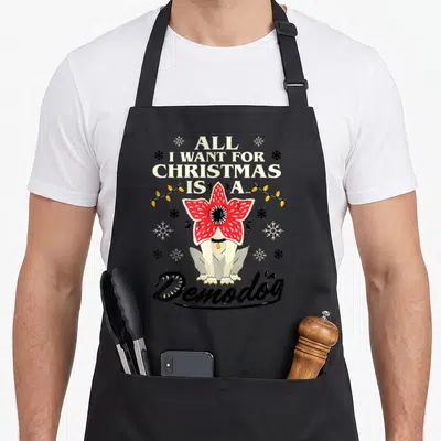 Tablier de cuisine noir avec illustration d'un Demodog transformé en fleur rouge sous des guirlandes lumineuses et des flocons de neige.