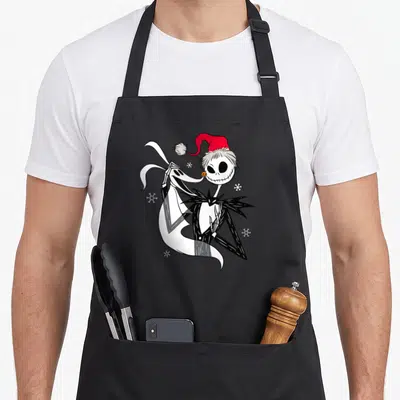 Tablier de cuisine noir avec un squelette en bonnet de Père Noël tenant un chien blanc entouré de flocons.
