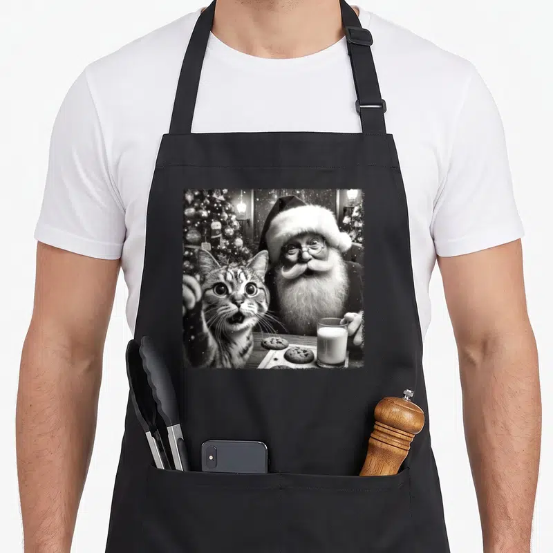 Tablier de cuisine noir avec une illustration monochrome d'un Père Noël et d'un chat en train de faire un selfie.