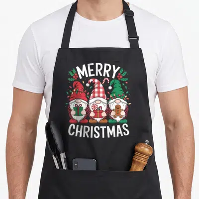 Tablier de cuisine noir avec trois nains de Noël festifs et l'inscription Merry Christmas en lettres blanches.