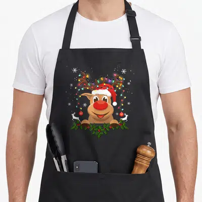 Tablier de cuisine noir avec renne de Noël portant un bonnet rouge et des guirlandes lumineuses autour des bois.