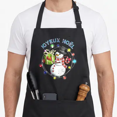 Tablier de cuisine noir avec un petit bonhomme de neige tenant un cadeau vert sous le texte JOYEUX NOËL et des lumières colorées.