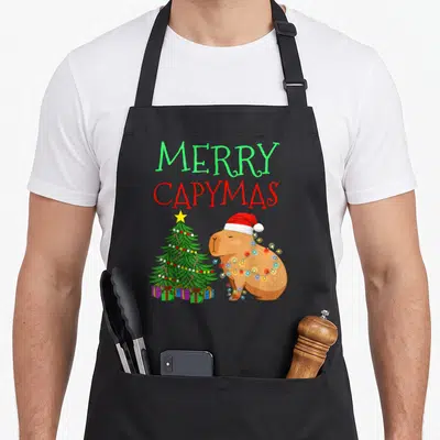 Tablier de cuisine noir avec illustration d'un capybara en bonnet de Père Noël près d'un sapin de Noël décoré.