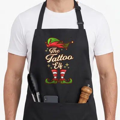 Tablier de cuisine noir avec illustration d'elfe aux bottes rayées et texte The Tattoo Elf entouré d'étoiles dorées.