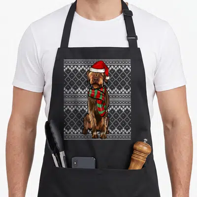 Tablier de cuisine noir avec un chien Griffon portant un bonnet de Noël et une écharpe verte et rouge sur fond de motifs géométriques blancs.