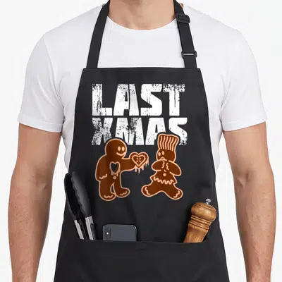 Tablier de cuisine noir avec inscription Last Xmas et deux bonshommes en pain d'épice échangeant un cœur.