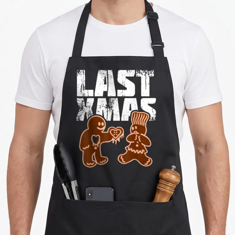 Tablier de cuisine noir avec inscription Last Xmas et deux bonshommes en pain d'épice échangeant un cœur.