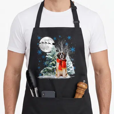 Tablier de cuisine noir avec un Saint Bernard en écharpe rouge devant une pleine lune et des flocons bleus.