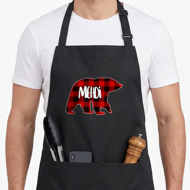 Tablier de cuisine noir avec un ours aux motifs buffalo plaid rouge et noir portant le prénom Mehdi en lettres blanches.
