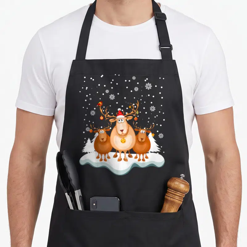 Tablier de cuisine noir avec trois élans marron, un bonnet rouge et des flocons de neige blancs sur fond blanc.