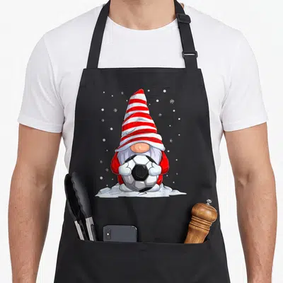Tablier de cuisine noir avec un nain de Noël portant une pointe rayée et tenant un ballon de football dans la neige.