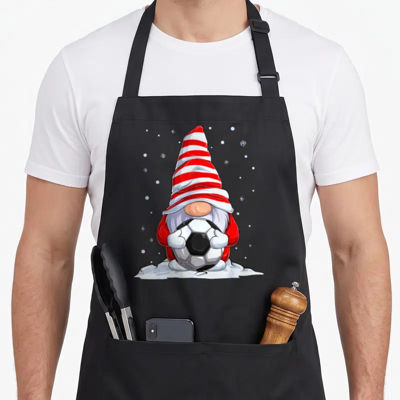 Tablier de cuisine noir avec un nain de Noël portant une pointe rayée et tenant un ballon de football dans la neige.