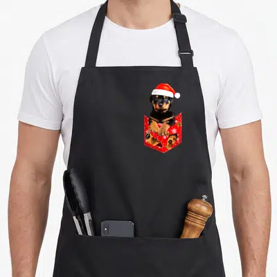 Tablier de cuisine noir avec un chiot Rottweiler portant un bonnet de Père Noël sortant d'une poche rouge à motifs festifs.