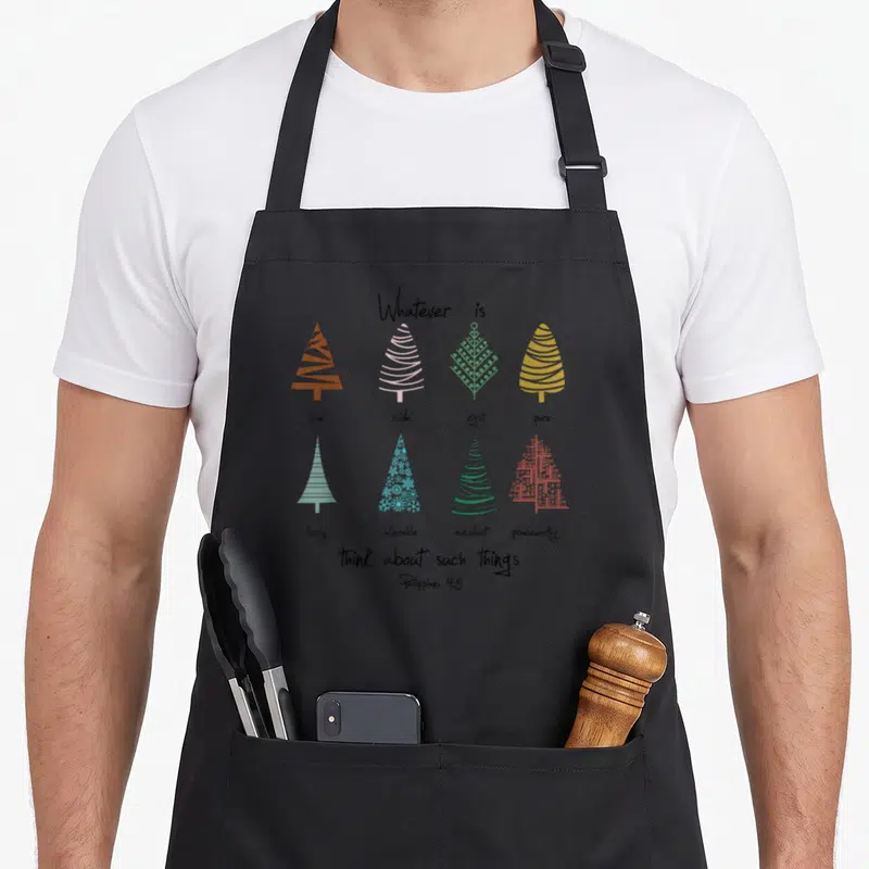 Tablier de cuisine noir avec sept arbres de Noël géométriques multicolores et un texte biblique en anglais centré sur le devant.
