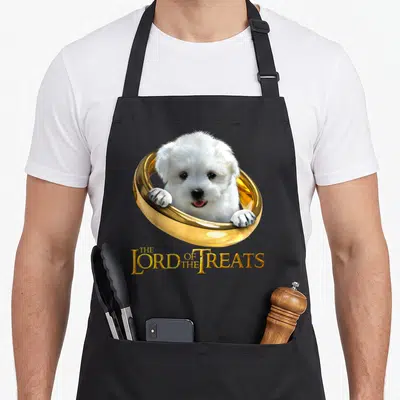 Tablier de cuisine noir avec un chiot maltais blanc dans une alliance dorée et le texte The Lord of the Treats en lettres capitales.