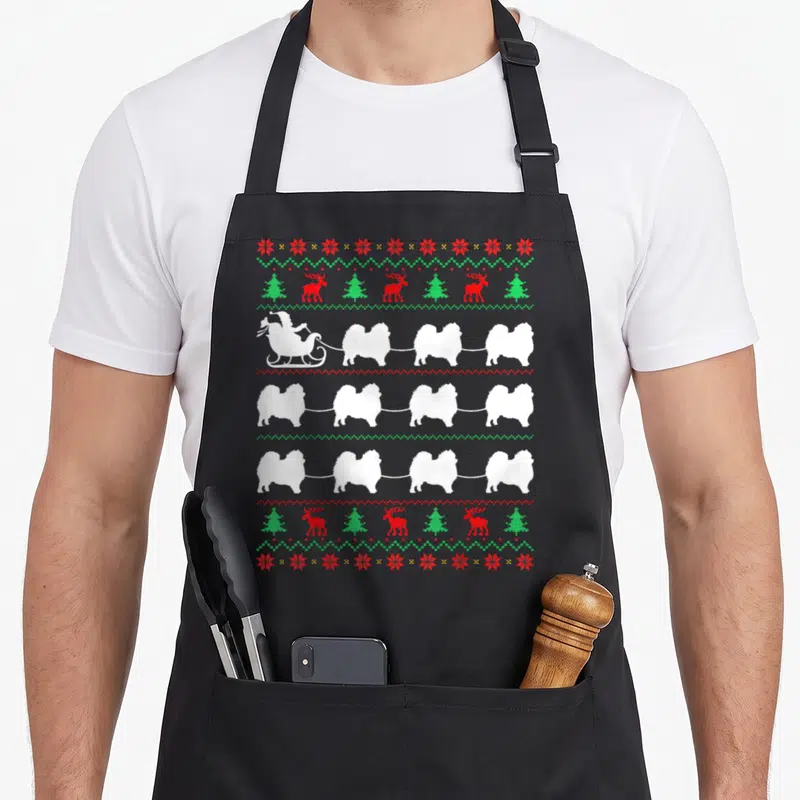 Tablier de cuisine noir avec motif de Pomeranians blancs tirant un traîneau de Père Noël sur fond gris uni.