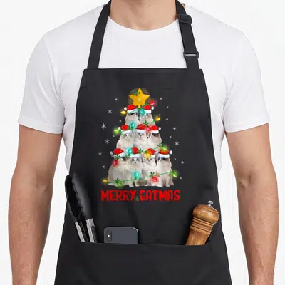 Tablier de cuisine noir avec six chats Ragdoll coiffés de bonnets rouges formant un sapin sous une étoile dorée et le texte Merry Catmas.