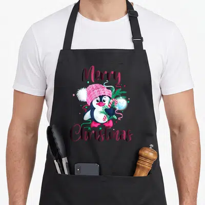 Tablier de cuisine noir avec pingouin en bonnet rose tenant une canne à sucre et l'inscription Merry Christmas.