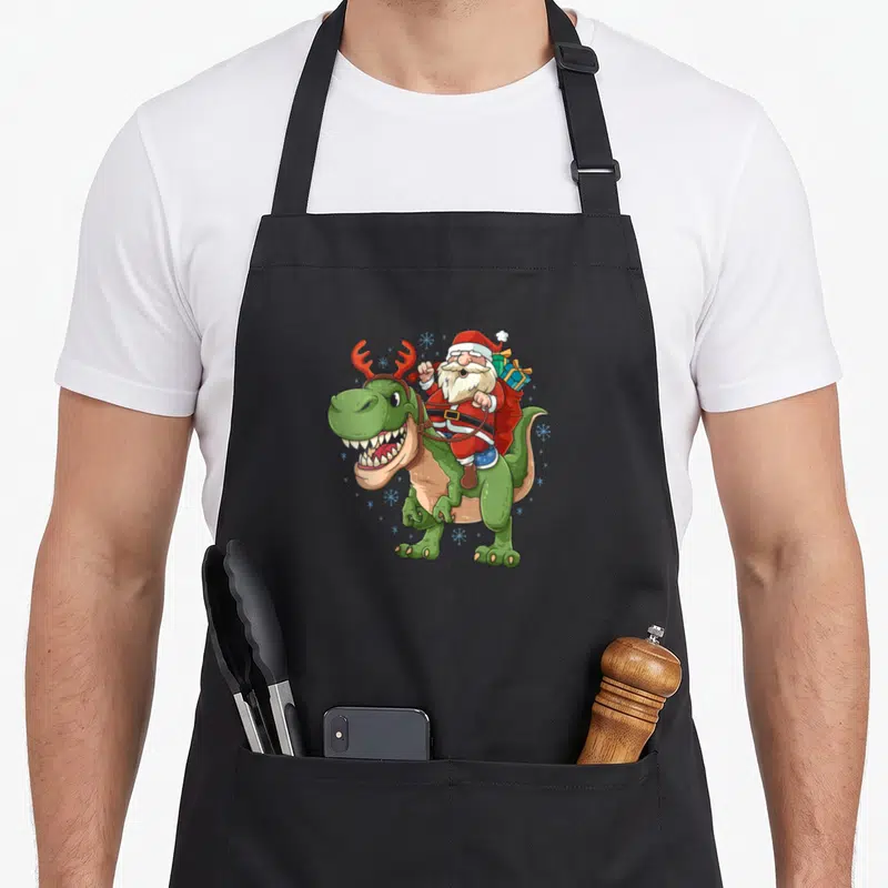 Tablier de cuisine noir avec illustration d'un Père Noël montant un T-Rex aux bois de renne et au sac de cadeaux.