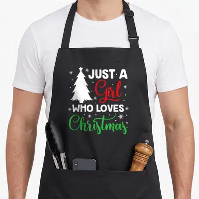 Tablier de cuisine noir avec un sapin blanc, des flocons et le texte Just A Girl Who Loves Christmas en lettres colorées.