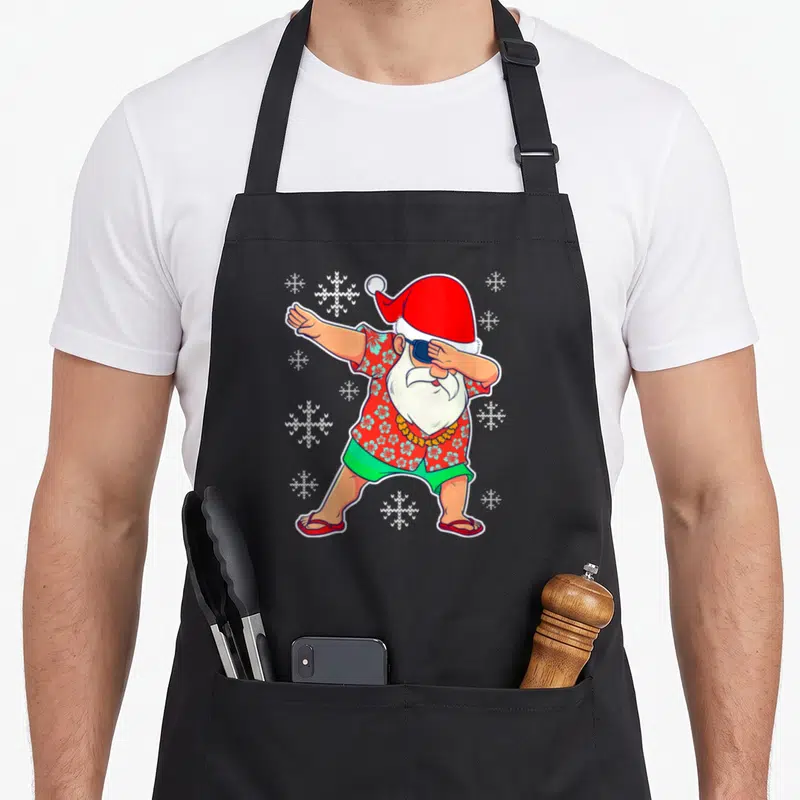 Tablier de cuisine noir avec illustration d'un Père Noël en tenue hawaïenne effectuant le dab entouré de flocons de neige.