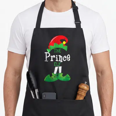 Tablier de cuisine noir avec illustration d'un elfe prince portant un chapeau rouge et des bottes vertes sur fond uni.