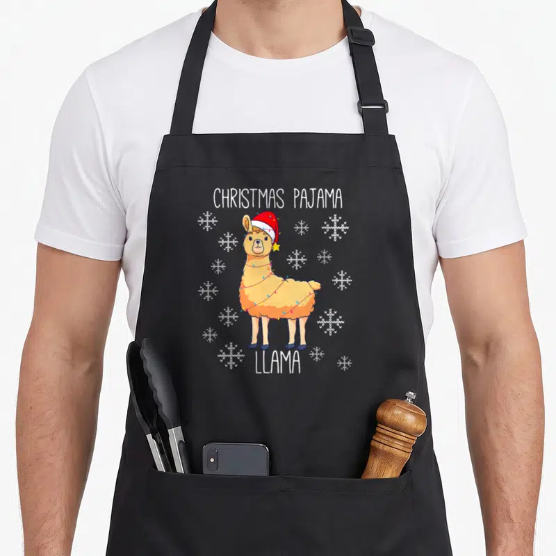 Tablier de cuisine noir avec illustration d'un lama portant un bonnet rouge et des lumières de Noël entouré de flocons blancs.