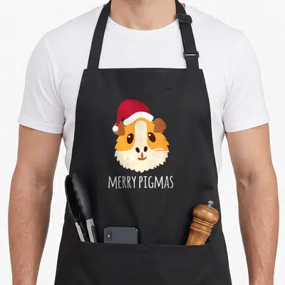 Tablier de cuisine noir avec tête de cochon d'inde portant un bonnet rouge et le texte Merry Pigmas en blanc.