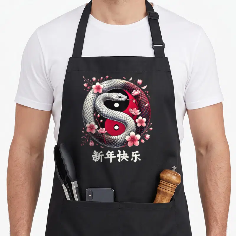 Tablier de cuisine noir avec un serpent entrelacé en yin yang entouré de fleurs roses et de calligraphie blanche.