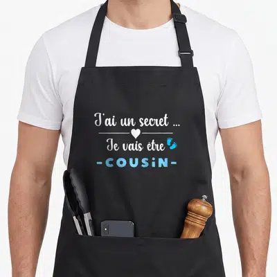 Tablier de cuisine noir avec texte blanc et bleu annonçant la venue d'un nouveau cousin en 2027.