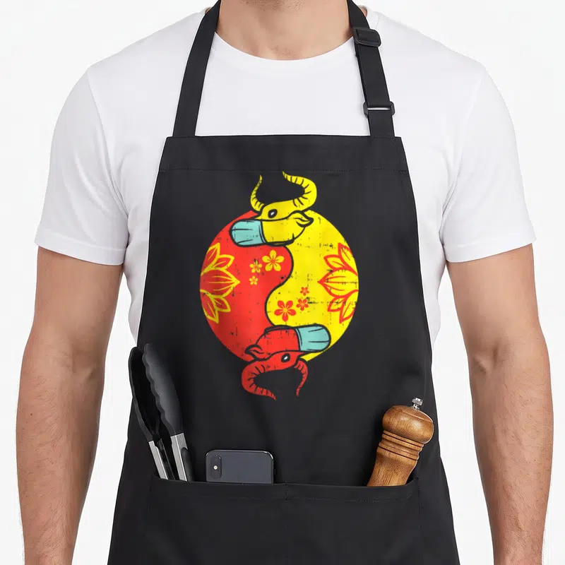 Tablier de cuisine noir avec un motif circulaire rouge et jaune représentant deux têtes de bœufs masqués en style Yin Yang.
