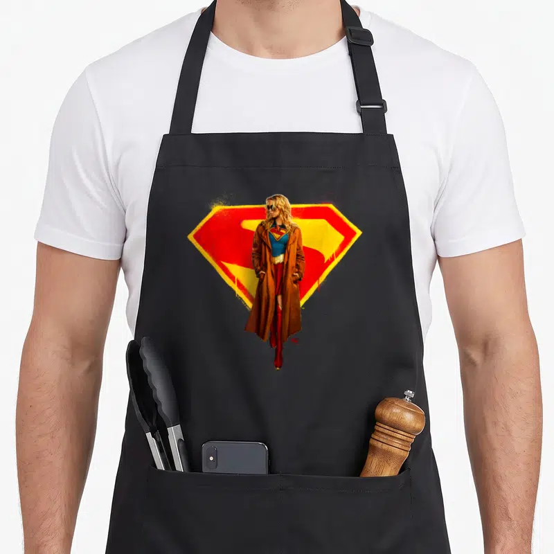 Tablier de cuisine noir avec Supergirl 2026 et Logo illustré en manteau brun et lunettes de soleil.