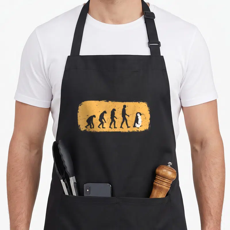Tablier de cuisine noir avec illustration évolutive menant à un pingouin debout sur fond orange texturé.