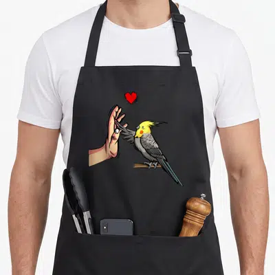 Tablier de cuisine noir avec illustration d'une calopsitte jaune et grise tendant son aile vers une main humaine sous un petit cœur rouge.