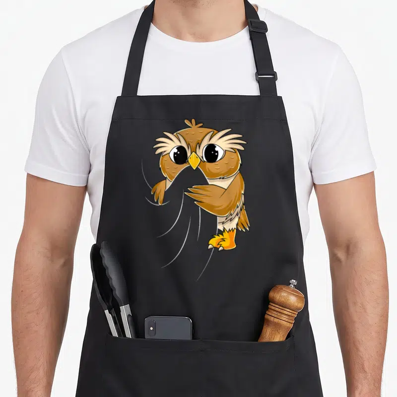 Tablier de cuisine noir avec un hibou marron aux grands yeux noirs et bec jaune tenant une serviette grise entre ses ailes.