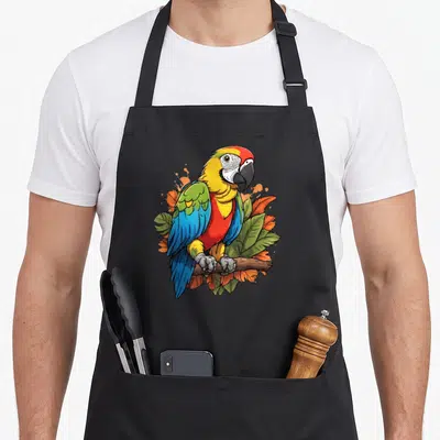 Tablier de cuisine noir avec un perroquet aux couleurs vives posé sur une branche au milieu de feuilles vertes et oranges.
