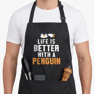 Tablier de cuisine noir avec un pingouin et le texte Life is Better with a Penguin en blanc et orange.