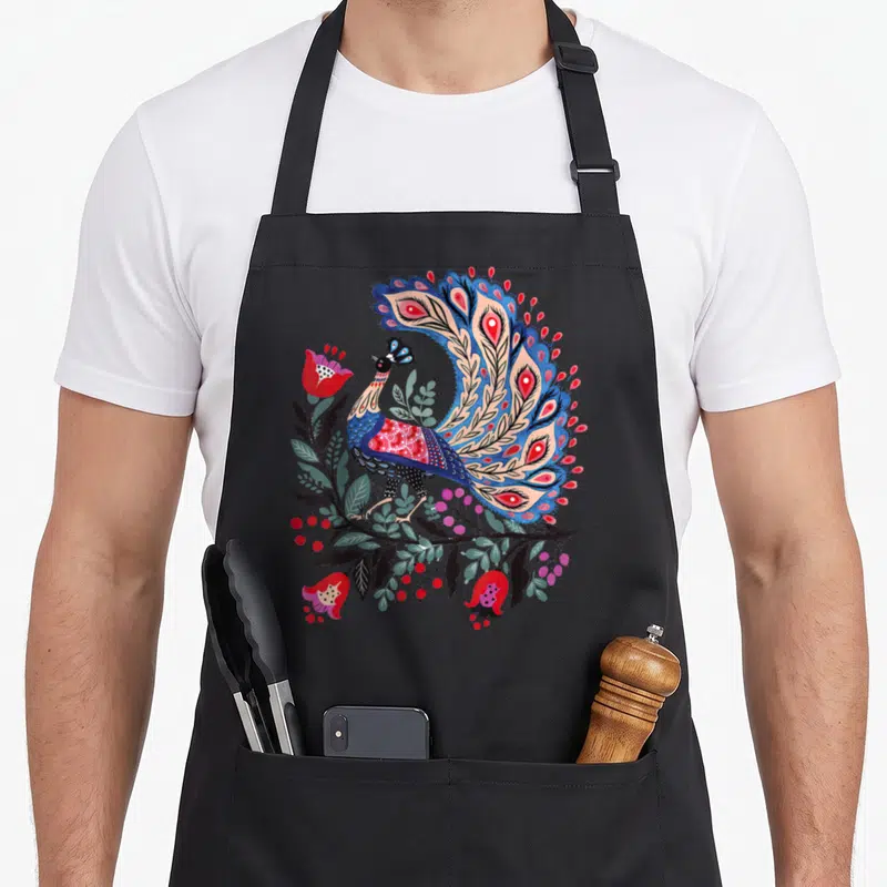 Tablier de cuisine noir avec un grand oiseau stylisé aux couleurs vives entouré de fleurs et de feuilles dans un style folklorique.