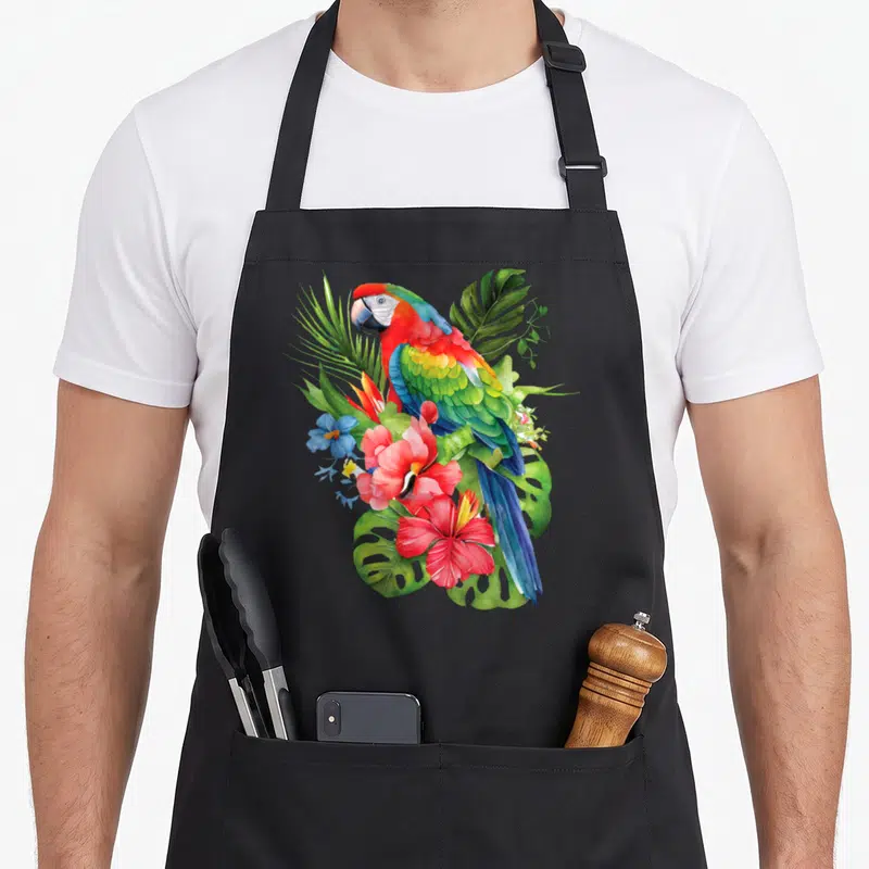 Tablier de cuisine noir avec un perroquet multicolore posé sur des fleurs roses et des feuilles de monstera vertes.
