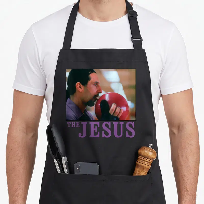 Tablier de cuisine noir avec une illustration d'un homme léchant une boule de bowling rouge et le texte The Jesus en violet en dessous.