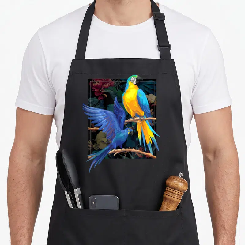Tablier de cuisine noir avec illustration de deux perroquets bleus et jaunes sur une branche florale tropicale.