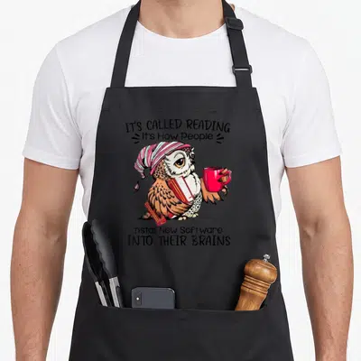 Tablier de cuisine noir avec hibou illustré tenant un livre rouge et une tasse rose, entouré de texte humoristique.