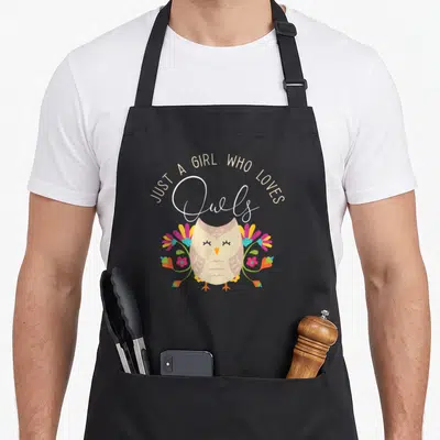 Tablier de cuisine noir avec un hibou stylisé entouré de fleurs colorées et du texte Just a girl who loves owls en écriture cursive.