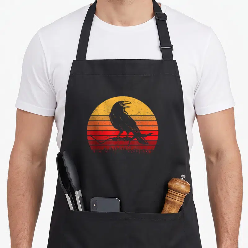 Tablier de cuisine noir avec illustration corbeau perchés sur branche devant soleil couchant rayé orange et rouge.