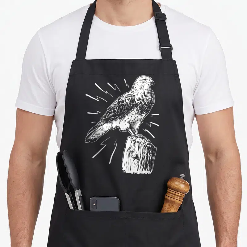 Tablier de cuisine noir avec illustration blanche d'un épervier perché sur un tronc d'arbre en style gravure.