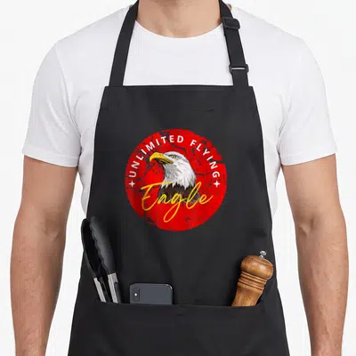 Tablier de cuisine noir avec illustration circulaire d'aigle blanc et texte UNLIMITED FLYING Eagle sur fond rouge texturé.