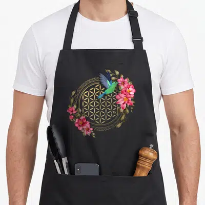 Tablier de cuisine noir avec un colibri coloré, une fleur de vie dorée et des fleurs roses dans un style mandala.